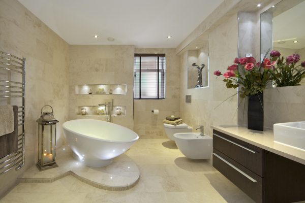 a modern bathroom in an expensive new home with a tear-drop shaped bath (full of water) sitting on a marble plinth. A bidet and WC are located near the window. A lantern with lit candles sits next to a large towel rail whilst a large bunch of red lilies sit on the cabinet to the right of the picture.